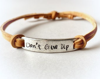 May include: A tan leather bracelet with a silver metal plate. The plate is engraved with the words "Don't Give Up" in a handwritten style font. The bracelet is tied with knots.