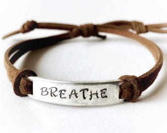 May include: A brown leather bracelet with a silver metal bar. The bar is stamped with the word "BREATHE" in a simple, sans-serif font. The bracelet is tied with a knot closure.