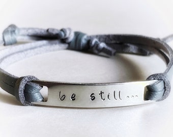 May include: A gray leather bracelet with a silver metal tag that reads "be still..."