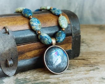 May include: A blue and green stone necklace with a round, dark gray pendant. The necklace is resting on a wooden box with metal accents.
