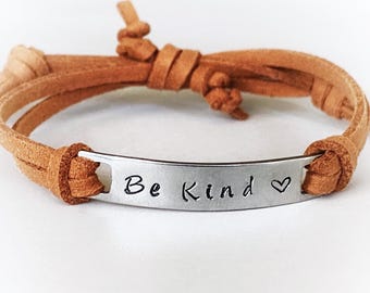 May include: A tan leather bracelet with a silver metal plate. The plate is engraved with the words "Be Kind" and a heart symbol. The bracelet is tied with a knot closure.
