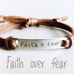 May include: A brown leather bracelet with a silver charm that reads "Faith > fear".