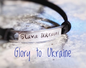 May include: A silver bracelet with the words "Slava Ukraini" engraved on a rectangular plate. The bracelet has a black cord band. The phrase "Glory to Ukraine" is written in blue script below the bracelet.