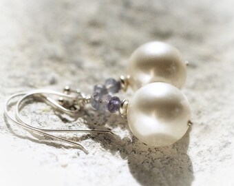 May include: A pair of drop earrings featuring large, round, off-white pearls suspended from silver-toned metal hooks. Each earring includes a small cluster of faceted, light purple beads positioned between the pearl and the silver wire hook.