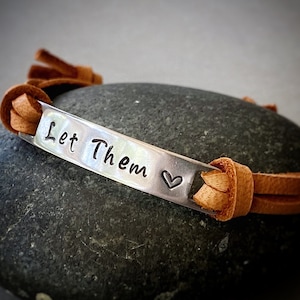 May include: A brown leather bracelet with a silver charm that says "Let Them" with a small heart symbol.