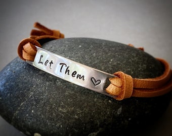 May include: A brown leather bracelet with a silver charm that says "Let Them" with a small heart symbol.