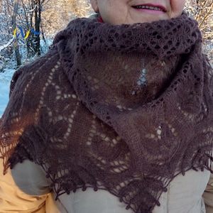 Może przedstawiać: Brązowa dziergana chusta z delikatnym ażurowym wzorem. Chusta jest udrapowana wokół szyi i ramion, w kształcie trójkąta. Krawędzie wykończone są falistym obszyciem. Tło przedstawia zaśnieżoną zimową scenerię.