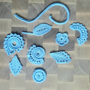May include: A collection of light blue crochet embellishments in various shapes, including leaves, flowers, and spiral designs. The handmade pieces are arranged on a light wood-grain surface, showcasing the intricate details of the yarn work.