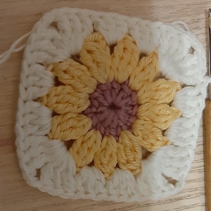 May include: A square crochet piece featuring a sunflower design. The center is a muted purple, surrounded by yellow petals, and framed by a white border. The yarn is thick, giving the piece a textured appearance. A crochet hook is visible on the right side.