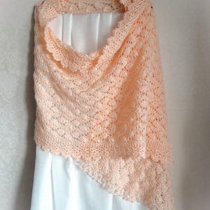 May include: A peach-colored crocheted shawl with a delicate lace pattern. The shawl is draped over a white hanger and a white fabric background.
