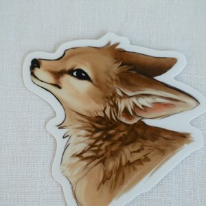 May include: A sticker featuring a detailed illustration of a fox's head, looking up with a soft expression. The fox has brown fur with a lighter shade on its face and ears. The background is white.