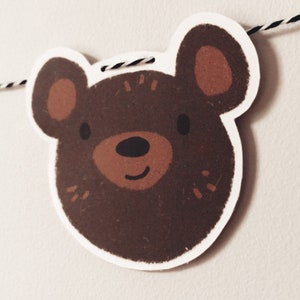 May include: A brown bear face with a white outline, brown ears, and a black nose. The bear is smiling and has a white background.