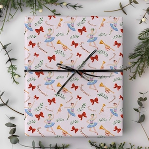 May include: Gift wrap with a repeating pattern of ballerinas in blue tutus, red bows, gold trumpets, and candy canes on a pale pink background. The wrapped gift is tied with a black ribbon.