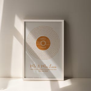 May include: A framed print featuring the song lyrics to "Perfect" by Ed Sheeran. The design includes a circular graphic with the song title and artist's name in a warm orange hue. The bottom of the print displays the names "Mr & Mrs Lucie" and a date.