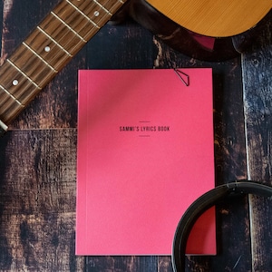 Puede incluir: Un libro de letras rosa con el título "SAMMI'S LYRICS BOOK" impreso en negro en la portada. Una guitarra, unos auriculares y un clip de papel son visibles en el fondo.