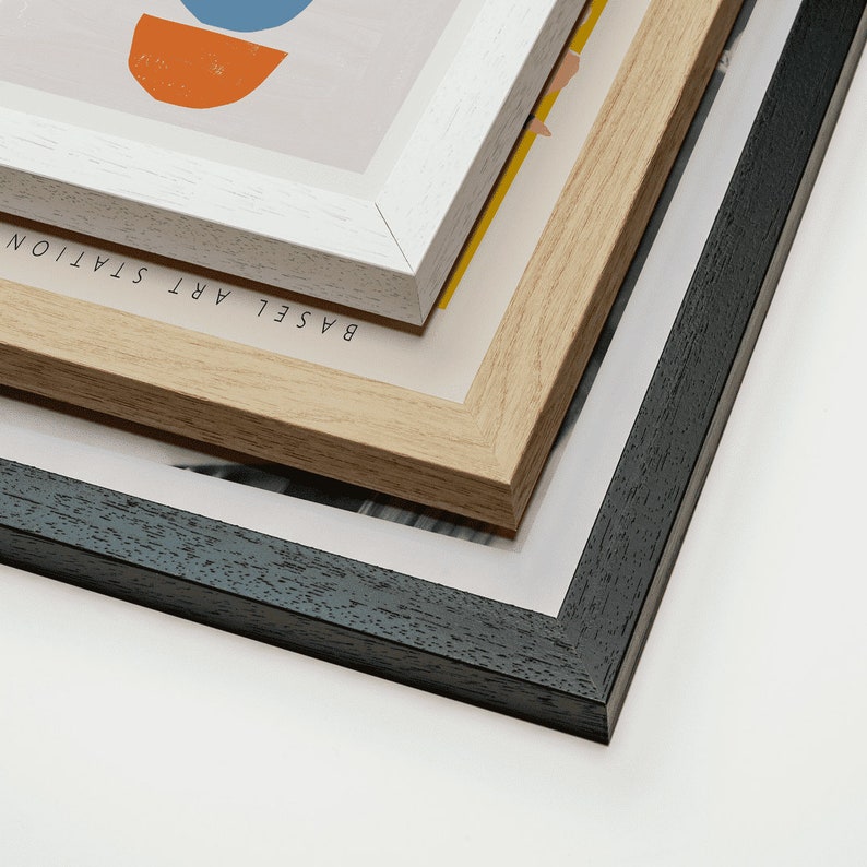 May include: Three picture frames stacked on top of each other. The top frame is white with the text "BASEL ART STATION". The middle frame is light brown wood. The bottom frame is black.
