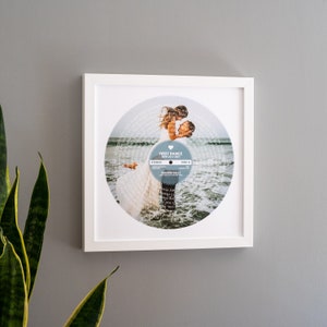 May include: White framed print of a wedding photo with the couple dancing on a beach. The photo is in the shape of a record with the lyrics of the song "Can't Take My Eyes Off You" by Frankie Valli printed around the edge. The text "First Dance 30th July 2017" is printed on the record.