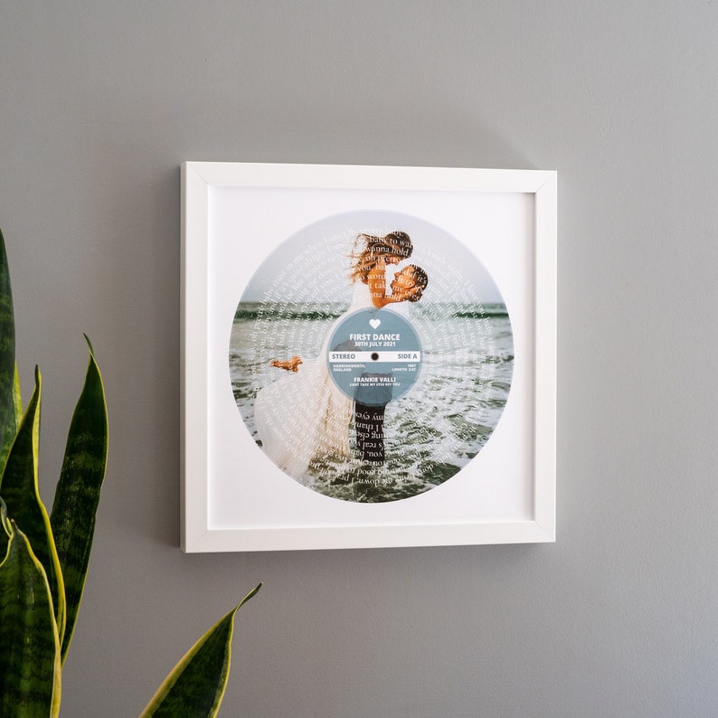 May include: White framed print of a wedding photo with the couple dancing on a beach. The photo is in the shape of a record with the lyrics of the song "Can't Take My Eyes Off You" by Frankie Valli printed around the edge. The text "First Dance 30th July 2017" is printed on the record.