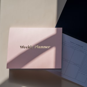 Op de afbeelding: Een roze weekplanner met gouden folielettering die "Weekly Planner" zegt. De planner is open op een wekelijkse overzichtspagina met een rasterindeling voor elke dag van de week.