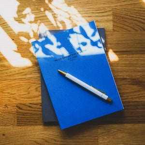 Peut inclure: Un carnet bleu avec la citation "You can make anything by writing." - C.S. Lewis imprimée sur la couverture. Un stylo blanc avec une pointe dorée est posé sur le carnet.