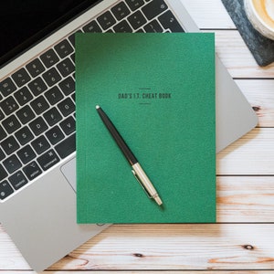 May include: A green notebook with the text "DAD'S I.T. CHEAT BOOK" on the cover. A black pen is lying on top of the notebook.