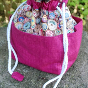May include: A pink drawstring bag with a white cord and a fabric lining featuring a pattern of colourful spools of thread.