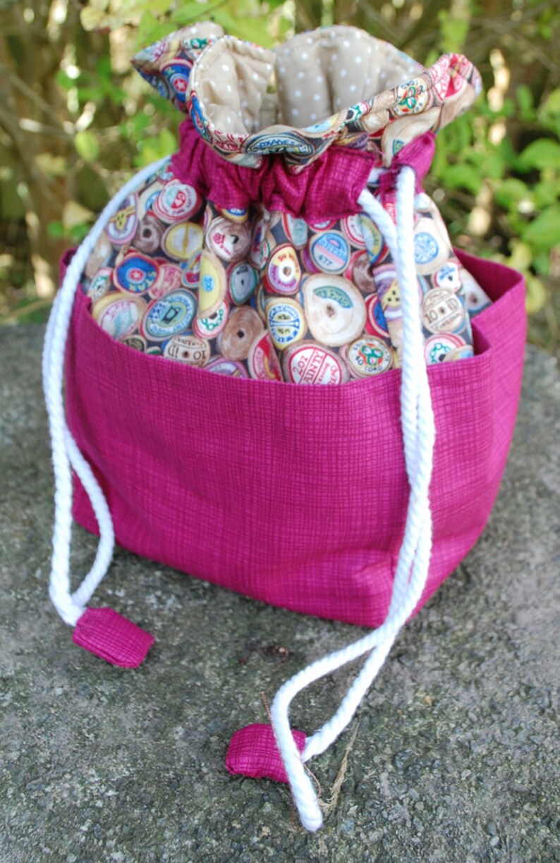 May include: A pink drawstring bag with a white cord and a fabric lining featuring a pattern of colourful spools of thread.