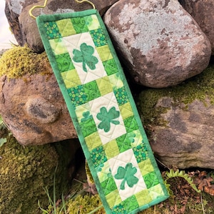 May include: A green and white quilted wall hanging with three shamrock appliques. The rectangular piece is made of various green patterned fabrics and features a gold-colored hanger at the top. The background includes moss-covered rocks.