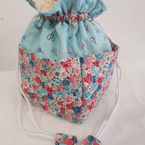 May include: A light blue drawstring bag with a button pattern on the bottom half and a light blue fabric with a pattern of scissors on the top half. The bag has white drawstrings with fabric ends.