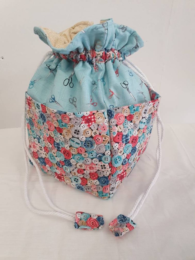 May include: A light blue drawstring bag with a button pattern on the bottom half and a light blue fabric with a pattern of scissors on the top half. The bag has white drawstrings with fabric ends.