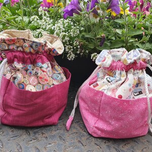 May include: Two drawstring bags with pink fabric and different patterns. The bag on the left has a cream fabric with a pattern of vintage spools of thread. The bag on the right has a cream fabric with a pattern of sewing tools.