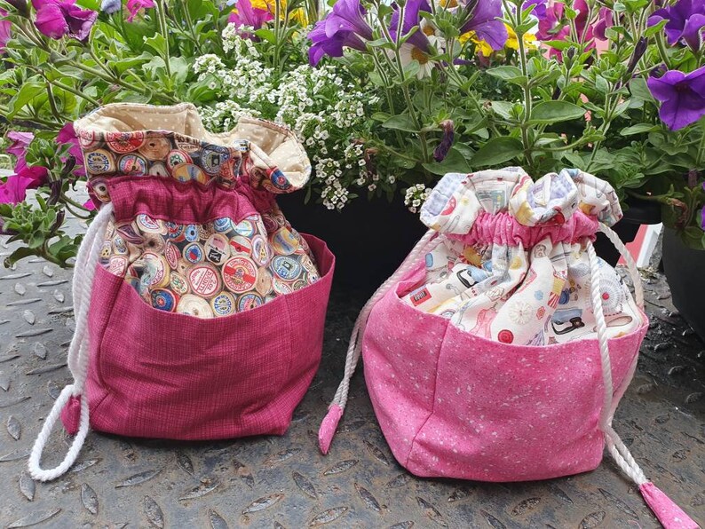 May include: Two drawstring bags with pink fabric and different patterns. The bag on the left has a cream fabric with a pattern of vintage spools of thread. The bag on the right has a cream fabric with a pattern of sewing tools.