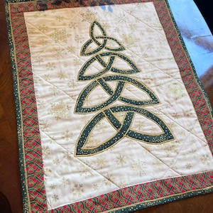 May include: A white quilted table runner with a green and gold Celtic knot design in the shape of a Christmas tree. The runner has a red and green border with gold accents.