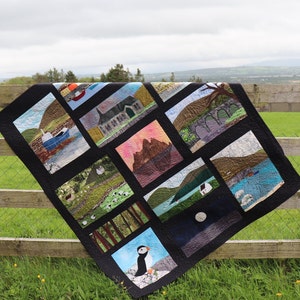 Könnte beinhalten: Eine schwarze Steppdecke mit 12 verschiedenen Quadraten, die verschiedene Landschaften und Szenen aus der irischen Landschaft zeigen. Die Steppdecke hängt an einem Holzzaun vor einem grünen Grasfeld.