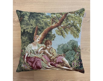 May include: A square throw pillow featuring a woven tapestry design of two figures sitting under a tree in a garden. The scene includes a person in a white and burgundy outfit and another in a gold and pink outfit, surrounded by green foliage.