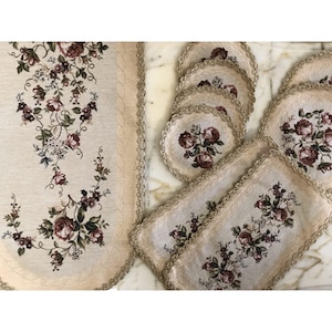 May include: A set of beige table linens with a floral pattern. The set includes a table runner and six placemats. The linens have a delicate lace trim.