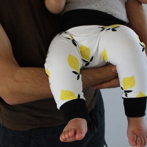 Può includere: Un bambino indossa leggings bianchi con stampa a limone e polsini neri. I leggings sono tenuti da un adulto.
