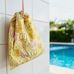 May include: A drawstring bag with a yellow background and a repeating pattern of orange and pink flip-flops. The bag is hanging on a white tiled wall with a red string. In the background is a swimming pool.