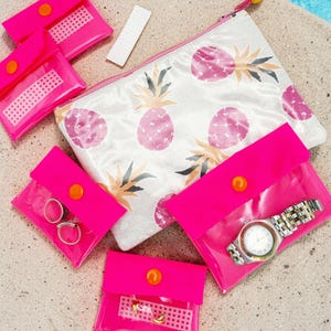 Può includere: Una collezione di accessori da viaggio, tra cui diverse piccole pochette rosa acceso con chiusure a scatto arancioni, un oggetto rettangolare bianco e una grande pochette con cerniera con un motivo ad ananas. Un orologio d'argento e anelli sono visibili.
