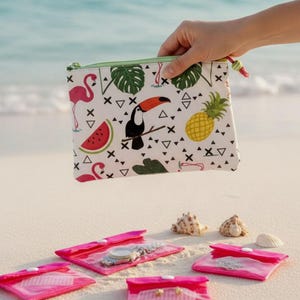 May include: A white zippered pouch with a tropical print featuring a toucan, flamingo, watermelon, and pineapple. Several pink travel pouches and seashells are on the sand. The pouch is held by a hand.