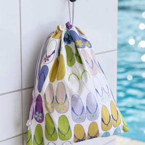 May include: A drawstring bag with a white background and a pattern of colorful flip-flops in shades of purple, green, yellow, and pink. The bag is hanging from a black hook on a white tiled wall, with a pool in the background.