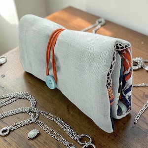 May include: A rolled-up, pale grey linen jewellery roll fastened with an orange cord and a blue button. The interior lining has a colourful floral pattern. Several silver necklaces are displayed on a wooden surface.