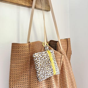 May include: A tan leather tote bag with a laser-cut design, hanging from a wooden hook. A small leopard print pouch with a yellow zipper is attached to the bag. The bag has long, light-colored straps.