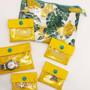 May include: A large zippered pouch with a banana and monstera leaf print, accompanied by five smaller yellow vinyl pouches. Each small pouch features a clear front window, a yellow flap, and a bright blue snap closure. One pouch contains a silver watch.