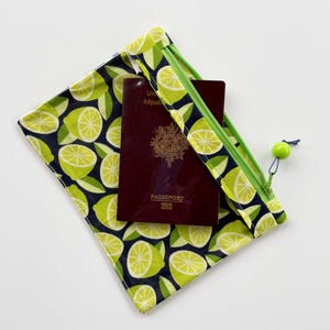 Peut inclure: Une pochette en tissu rectangulaire avec un fond bleu marine foncé orné d'un motif répétitif de tranches de citron vert et de feuilles. La pochette possède une fermeture éclair vert citron avec une perle assortie et contient un passeport bordeaux.
