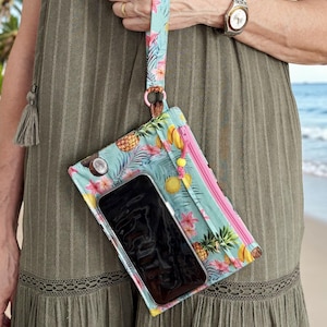 May include: A turquoise wristlet featuring a tropical fruit and floral design with pineapples, lemons, and pink flowers. The wrist strap and zip are pink. A smartphone is visible inside the clear pocket. The wristlet is held by a person.
