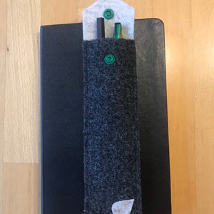Könnte beinhalten: Ein grauer Filz-Stifthalter mit einem weißen Blatt-Design und grünen Knöpfen. Der Halter ist an einem schwarzen Notizbuch befestigt.