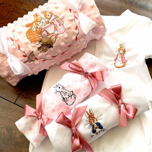 May include: A set of three baby blankets featuring Peter Rabbit and friends. The blankets are pink and white with embroidered designs. The blankets are tied with pink satin ribbons.