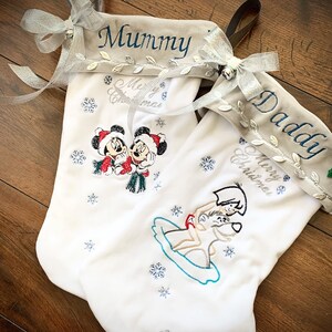 May include: Two white Christmas stockings with embroidered designs. One stocking has Mickey and Minnie Mouse, the other has a dog and a person. The top of each stocking is gray with silver trim and the words "Mummy" and "Daddy" embroidered on them.