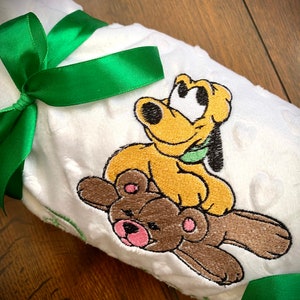 Puede incluir: Una manta de felpa blanca con una cinta verde atada alrededor. La manta presenta un diseño bordado de un perro de dibujos animados amarillo sentado sobre un oso de peluche marrón.